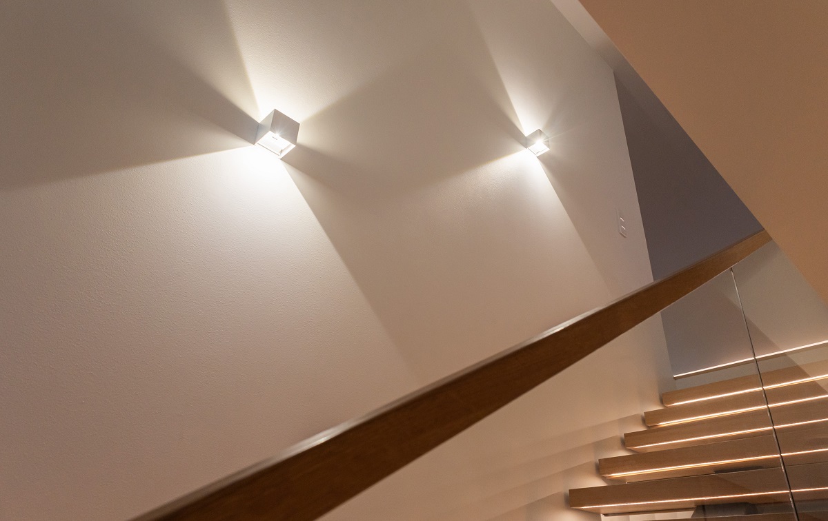 A modern staircase with wooden steps and a glass railing and LED wall lights creating light patterns on the wall. 
