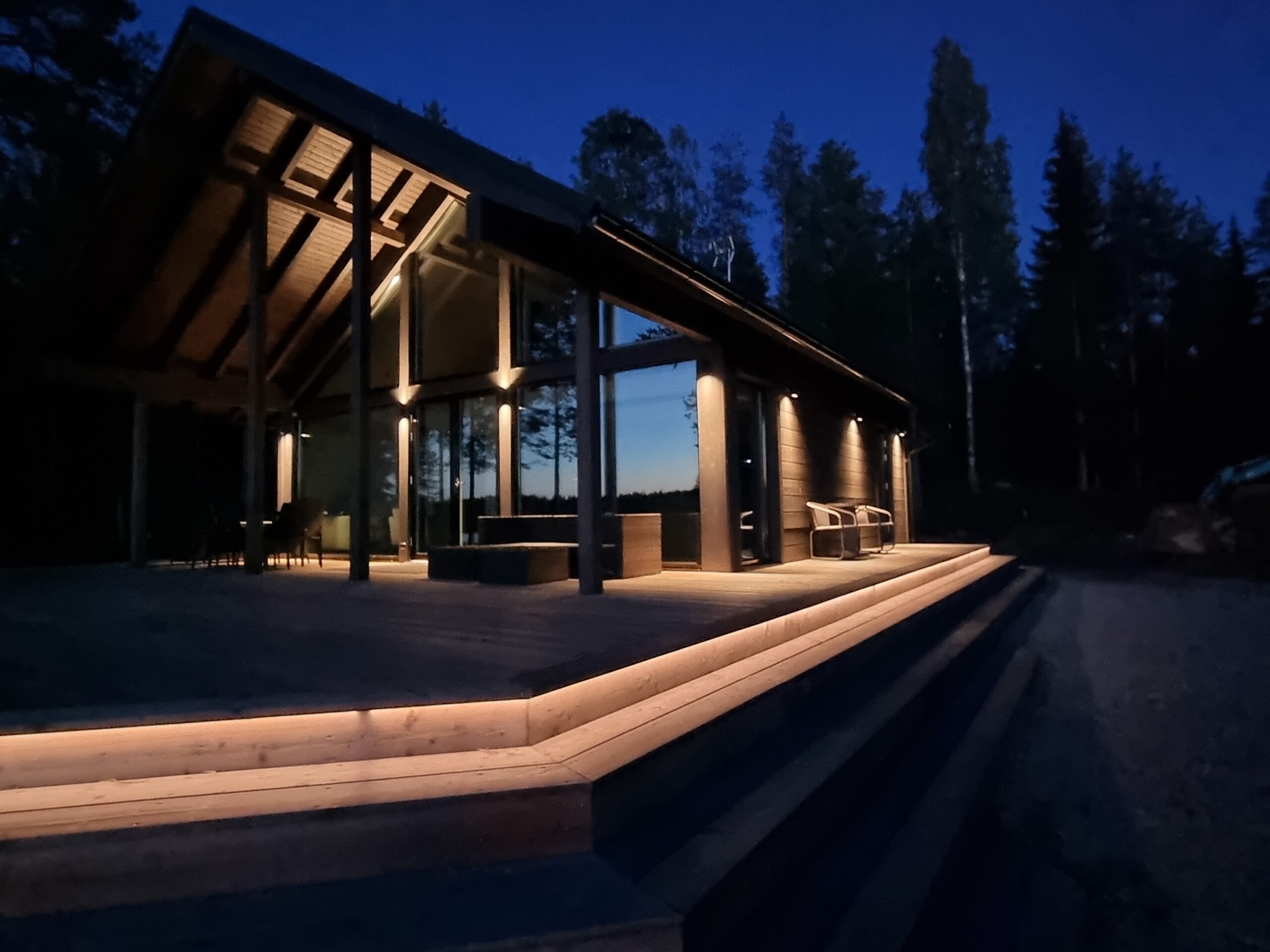 Nykyaikainen hirsimökki, jossa on suuret ikkunat ja ulkoportaat, kylpee lämpimässä tunnelmavalossa LedStore.fi:n laadukkaan ulkovalaistuksen ja led-valonauhojen mukana. Tyylikkäät led-valaisimet korostavat mökin arkkitehtuuria sekä ympäriä luontoa, luoden kutsuvan ilmeen iltahämärässä. - LedStore LED strip lights can be installed over long distances as a continuous line. In this example, the steps surrounding the terrace are illuminated so that an LED strip is placed under the first step. The contrast created by the light clearly indicates where the first step begins.