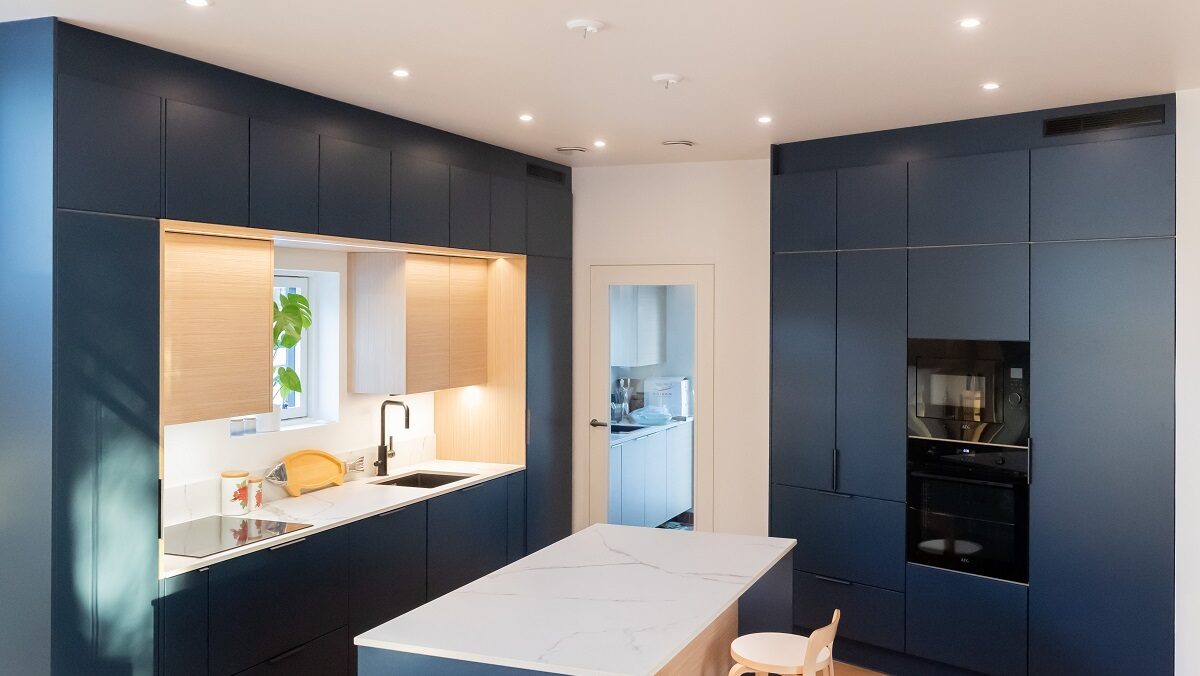 Moderni keittiö, jossa on tummansiniset kaapit, puuosat, marmorisaari ja sisäänrakennetut laitteet, jotka on valaistu energiatehokkaalla LED-valaistuksella.