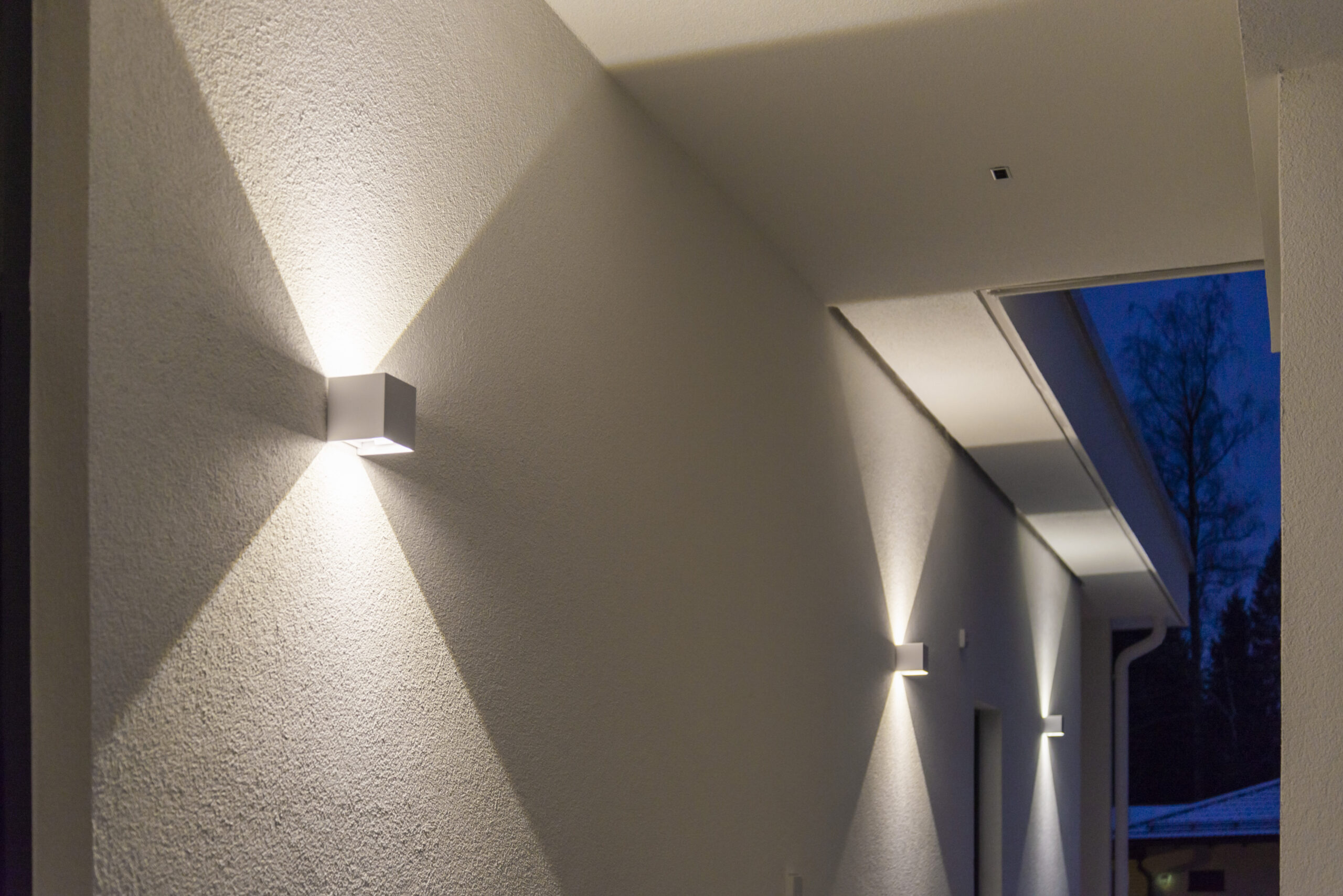 Modernit seinävalaisimet valaisevat vaatisti valkoisen ulkokäytävän modernin talon yhteydessä, luoden näyttävät X-kuvion valokeilat. Tämä on hienosta esimerkistä LedStoren led-pihavalaist, joka tuo ulkotiloihin toimivuutta eleganttia ilmettä. - LedStore Cube-shaped white LED wall lights illuminate both upward and downward, creating a sharp light pattern on the wall.