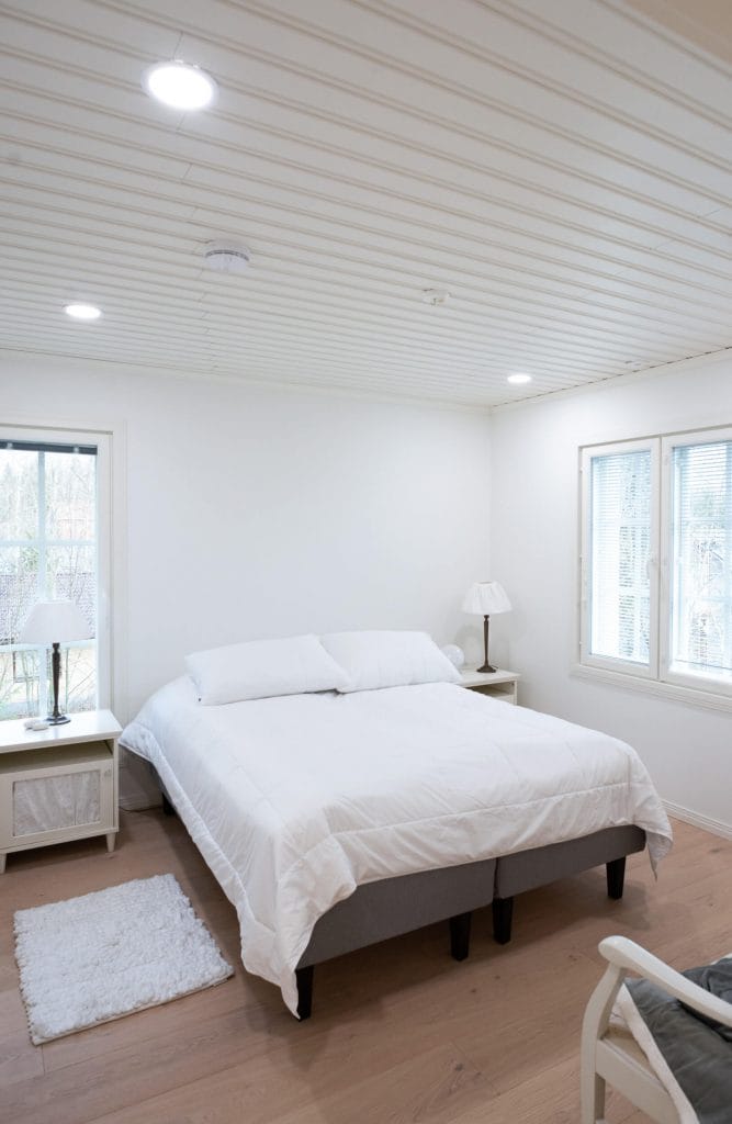 Viihtyisä makuuhuone, jossa valkoinen sänky, puulattia, yöpöydät lampuilla; upotettu LED-valaistus kattoon; luonnonvaloa kahdesta ikkunasta.