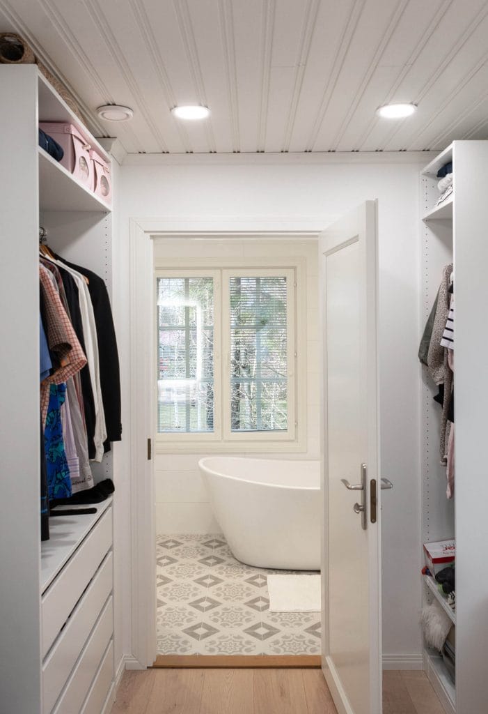 Avokaappi, joka johtaa moderniin kylpyhuoneeseen, jossa on valkoinen vapaasti seisova amme, iso ikkuna, kuviolliset lattialaatat ja upotetut LED-kattovalot.