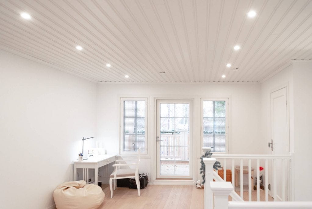 Minimalistinen huone, jossa upotettu LED-valaistus, valkoiset seinät, kirjoituspöytä, säkkipussi, pieni kasvi; lasiovi/ikkunat parvekkeelle; luonnonvaloa ja puulattiaa.