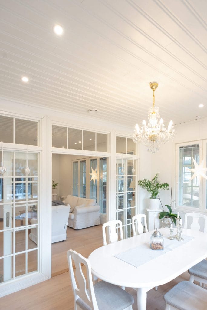 Ruokailutila, jossa on valkoinen pöytä ja tuolit, valaistu LED-kattokruunulla. Lasiovet johtavat olohuoneeseen, jossa on valkoiset huonekalut ja vihreitä ruukkukasveja.