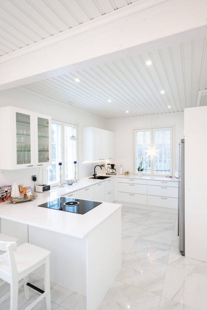 Moderni keittiö, jossa valkoiset kaapit, marmorilattia, saarekeliesi; LED-valaistus kirkastaa tilaa, kun luonnonvaloa tulvii sisään suurista ikkunoista.