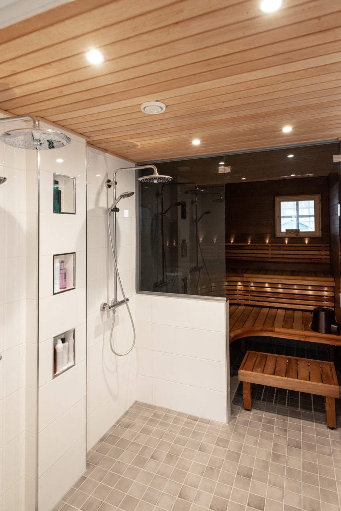 Moderni saunasuihku puisella katolla, kaksi sadesuihkua, LED-valaistus; lasiseinäisessä saunassa on puiset lauteet ja ikkunanäkymä.