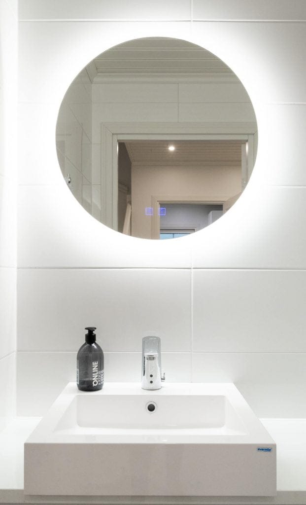 Minimalistinen moderni kylpyhuone, jossa on LED-taustavalaistu pyöreä peili, valkoinen neliön muotoinen pesuallas ja musta saippua-annostelija tyylikkääseen ja eleganttiin muotoiluun.