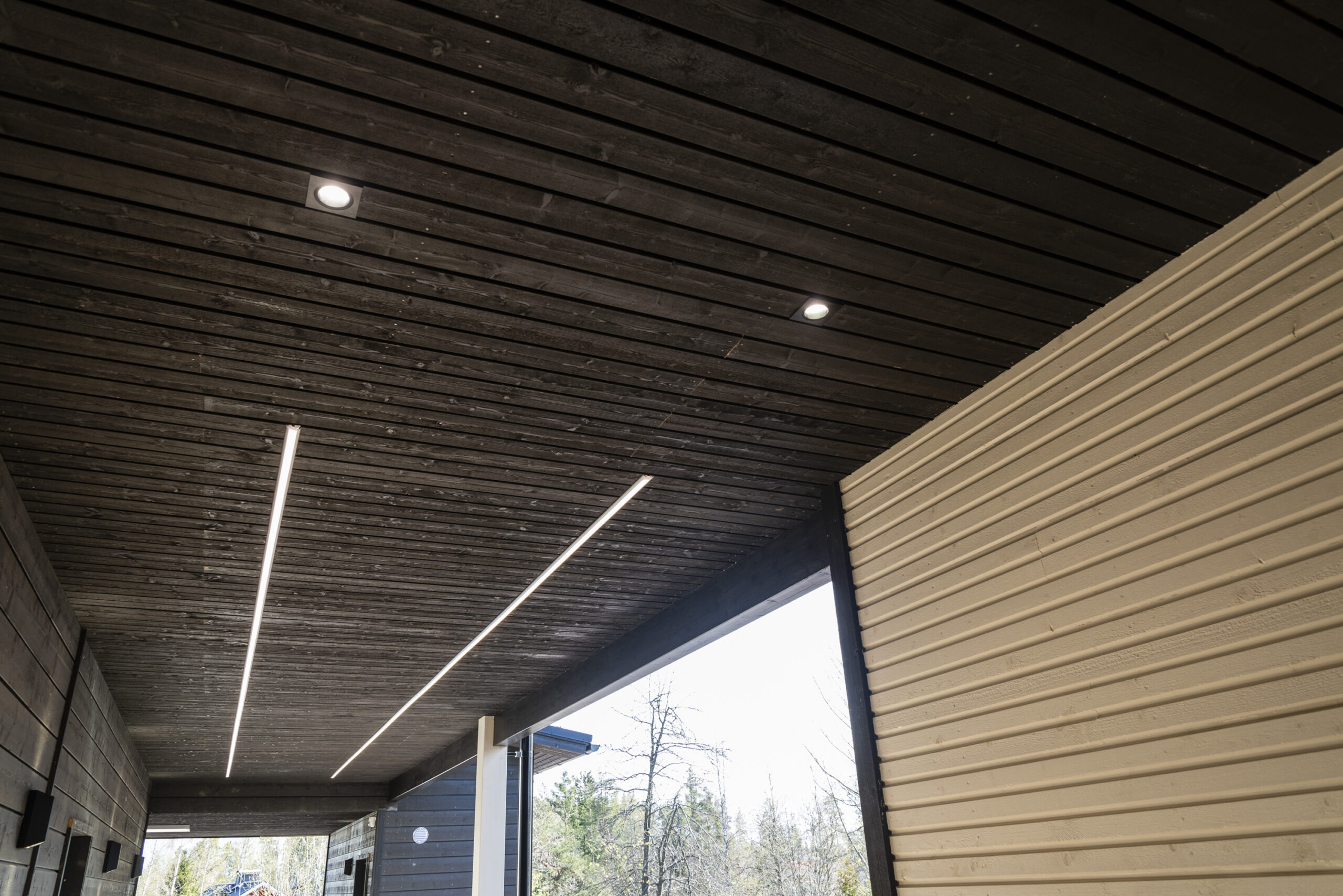 Puisessa katossa upotetut LED-alasvalot sekä LedStore.fi:n LED-pihavalonauhat valaisevat modernin talon ulkotilaa, jossa yhdistyy vaaleat ja tummat julkisivupinnat. - LedStore Along the entrance walkway, two long LED strip lights run parallel to light the path. In addition, two recessed LED spotlights and wall-mounted lights on the side enhance the overall illumination.