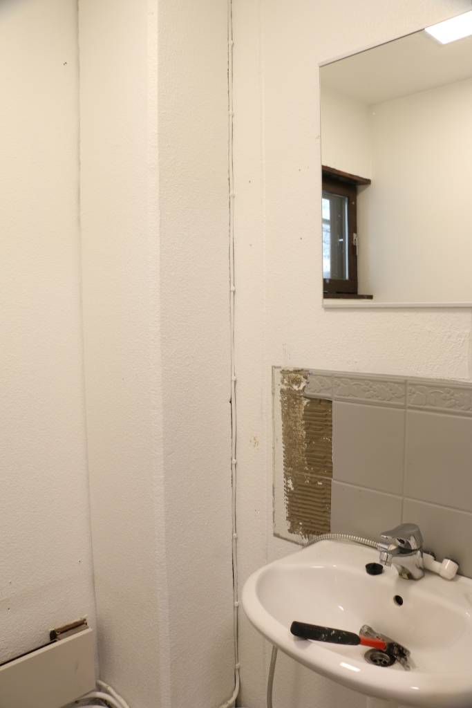 Before image for blog 1024px - LedStore A small bathroom with a LED-framed mirror above a white washbasin, silver faucet, and a visible cable; a black and red wrench on the washbasin. Partially removed tiles reveal the wall behind.