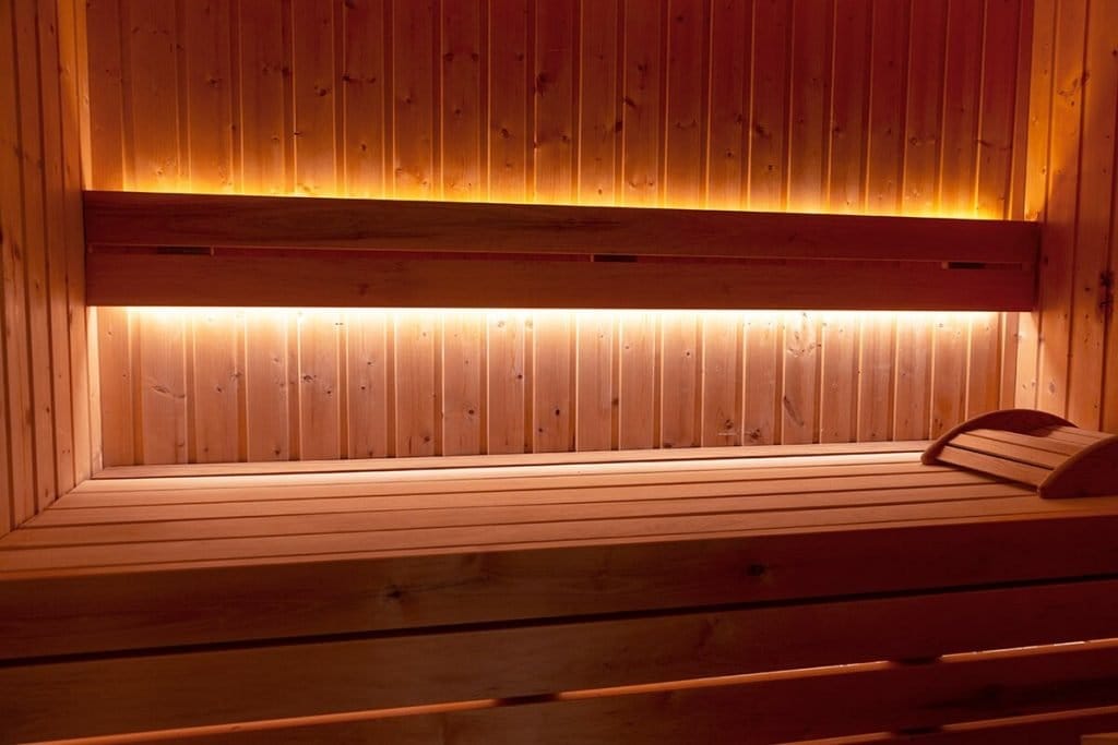 Wooden sauna interior featuring warm ambient LED lighting, wooden bench with headrest, and paneled walls and ceiling highlighting natural wood tones.