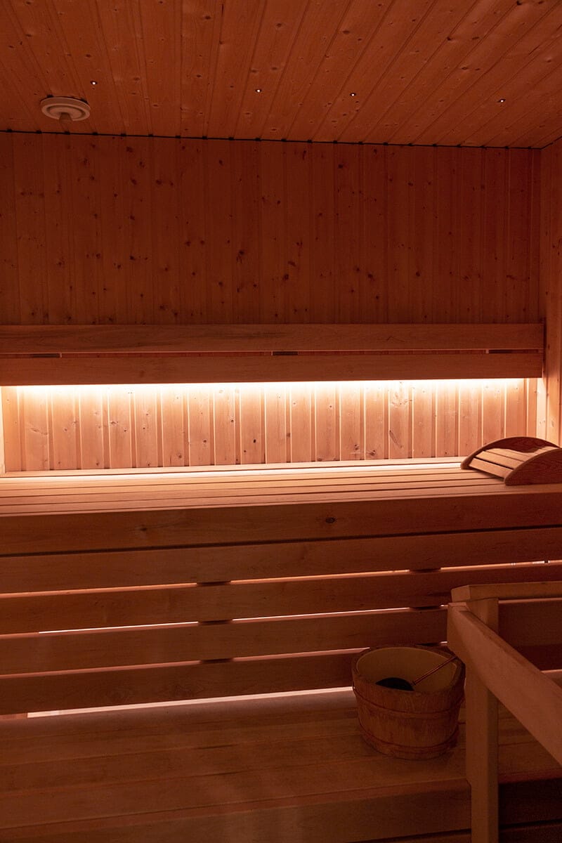Cozy wooden sauna interior with soft LED lighting, featuring light wood benches and walls, a sauna bucket on the bench, and a scoop creating warmth.