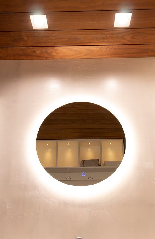 Circular mirror with LED border on a wooden wall, ceiling lights above, reflecting shelves lit by spotlights for modern LED lighting ambiance.
