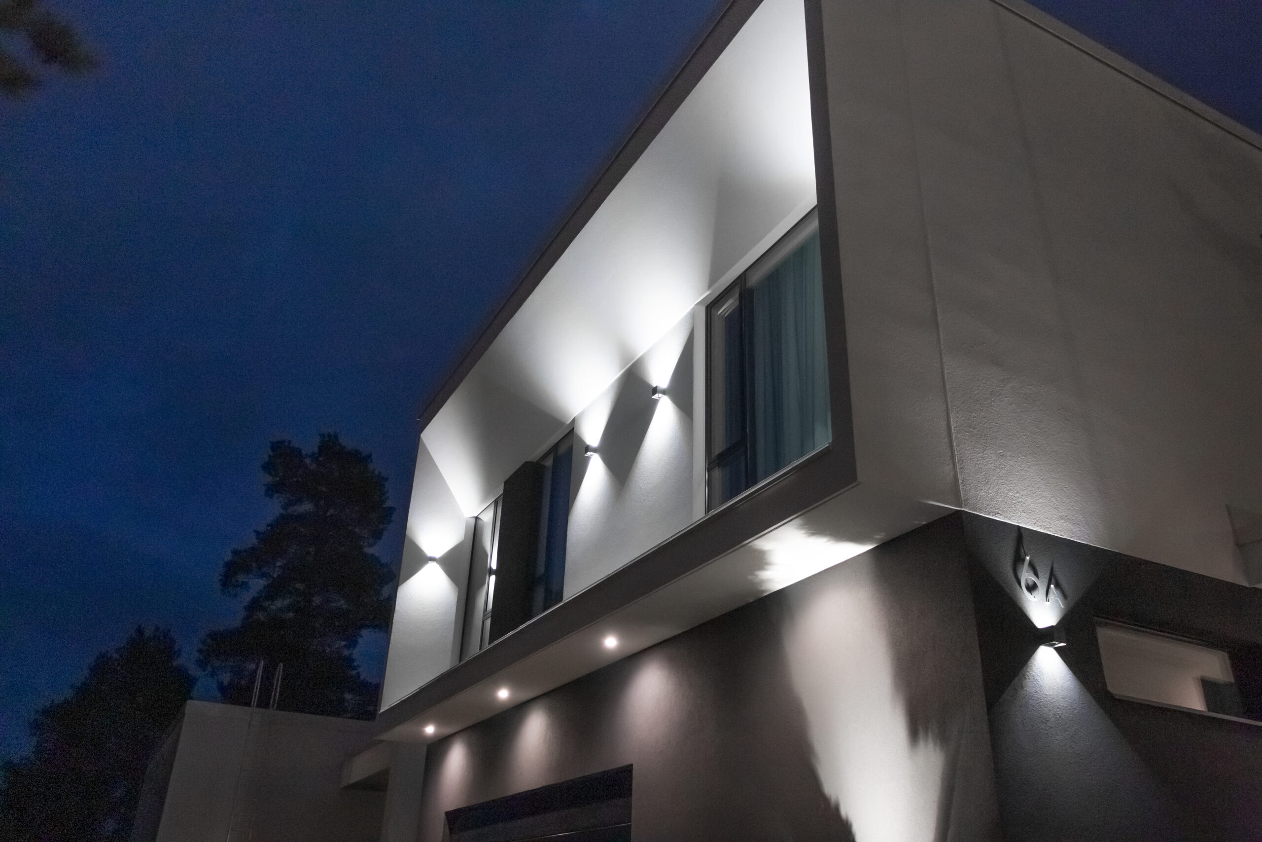 Moderni talo iltahämärässä – kulmikas arkkitehtuuri ja LedStore.fi:n laadukkaat LED-pihavalaisimet tuovat julkisivun näyttävästi esiin. - LedStore The facade of the house illuminated with wall lights and spotlights.