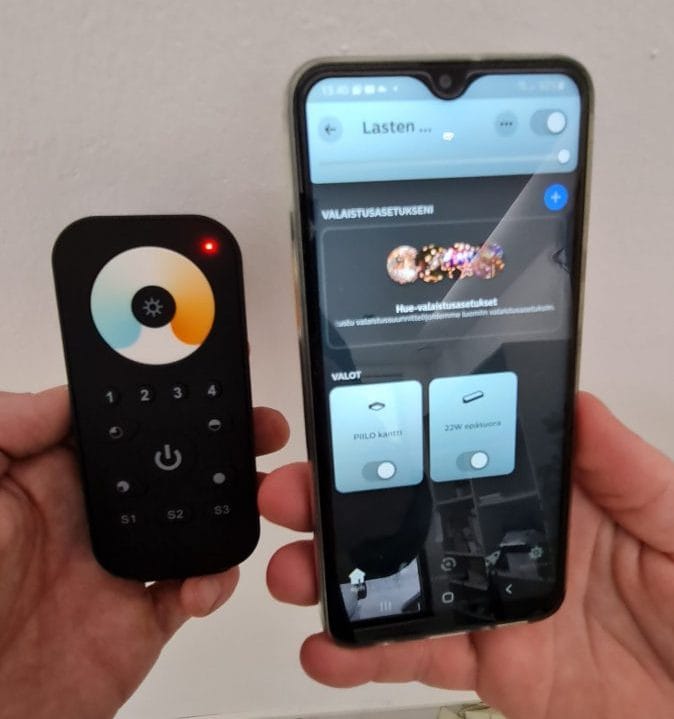 Käytetään himmennyksessä ja värilämpötilasäädössä mobiilissa ja kaukosäätimellä - LedStore Henkilö pitää kädessään älypuhelinta, jossa on LedStore.fi:n LED-valaistuksen ohjaussovelluksen käyttöliittymä, sekä kaukosäädintä, jossa on painikkeet ja väripyörä. Sovelluksen käyttöliittymässä on kaksi liukusäädintä ja yläpuolella väripaletti.