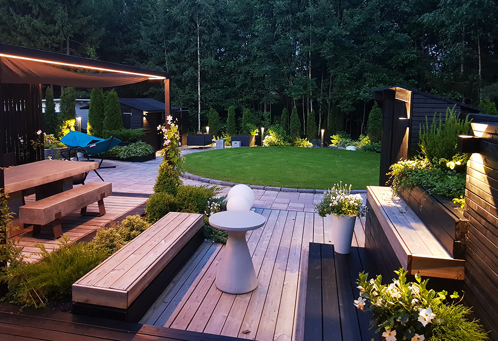 Nykyaikaisen talon modernilla takapihalla puiset viisit ja pyöreä pöytä kutsuvat viihtymään runsaan vehreyden keskelle. Tunnelmallinen LedStore.fi:n LED-pihavalaistus viimeistelee pihan, luoden täydelliset puitteet rentouttaviin kesäiltoihin. - LedStore An LED strip installed above the outdoor dining table provides overhead lighting, while wall lights on the right-hand wall illuminate in two directions.