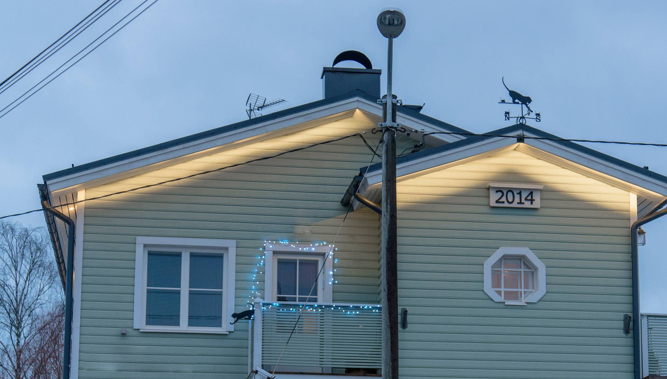 Vaaleanvihreä puutalo, jonka julkisivulla on vuosiluku "2014". Ulkovalaistuksessa tunnelmanauhat LedStore.fi:n ulkokäyttöön sopivat valosarjat sekä parvekkeella led-. Katolla koristuksena kissakuvioinen tuuliviiri. - LedStore LED strips used in outdoor lighting at the end of a pitched-roof house in two different configurations.