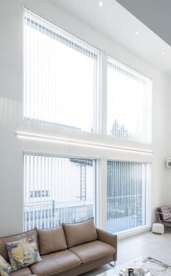 Led profile in a high living room - LedStore Modern living room with white vertical blinds, tan leather sofa and patterned pillows; soft LED lighting brightens walls and flooring.