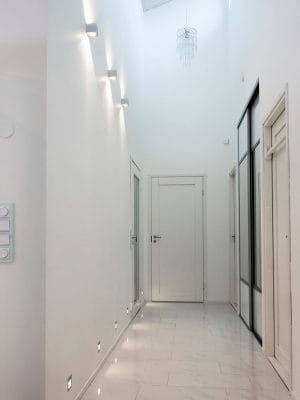 Decorating a corridor with lamps - LedStore Modern hallway with LED wall sconces, white walls, glossy floor, high ceiling light fixture, and multiple doors. Minimalist design.