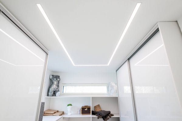 Wardrobe flush profile - LedStore Minimalist room featuring modern LED recessed ceiling lights, illuminated white shelves with vases and sculptures, sliding doors, and a horizontal window.