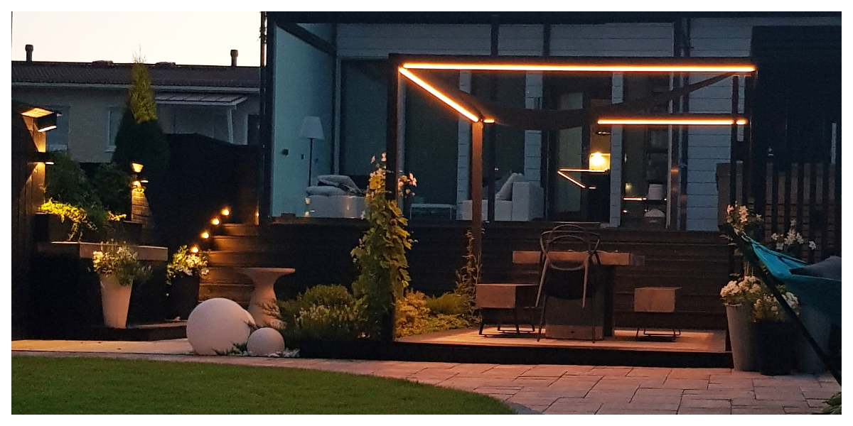 Nykyaikainen takapihan patio, jossa pergolaaa upea LED-tunnelmavalaistus. Puuporras ja tunnelmalliset ruukkukasvit. Modernin talon pihaan sijoittuva kokonaisuus tarjoaa täydelliset puitteet iltahetkiin LedStore.fi:n laadukkaalla led-valaistuksella. - LedStore Backyard patio with a pergola lit with LED strip. A wooden staircase illuminated from the side.