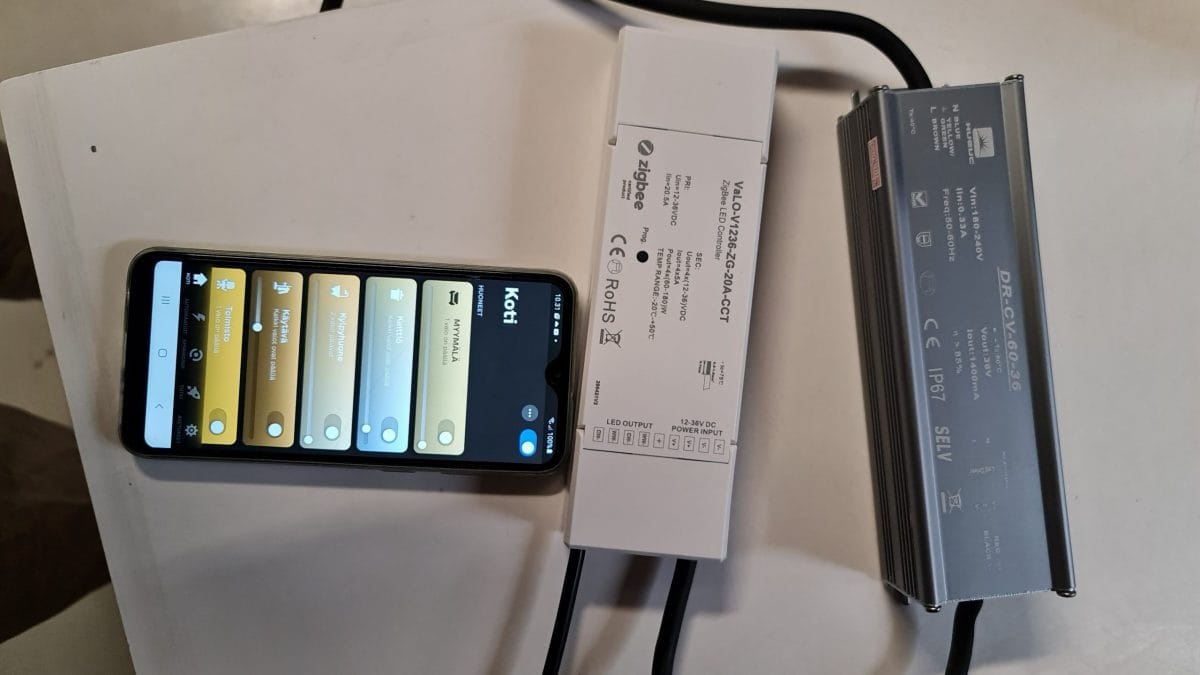 Muuntaja ZG vastaanotin CCT ja Hue applikaatio mobiilissa - LedStore Älypuhelin, jossa on auki kodin automaatiosovellus, sijaitsee kahden LED-valaistuksella varustetun elektronisen laitteen vieressä. Laitteissa on etiketit, joissa on yksityiskohtaiset tekniset tiedot, ja puhelimen näytöllä näkyy erilaisia valaistuksen säätömahdollisuuksia.