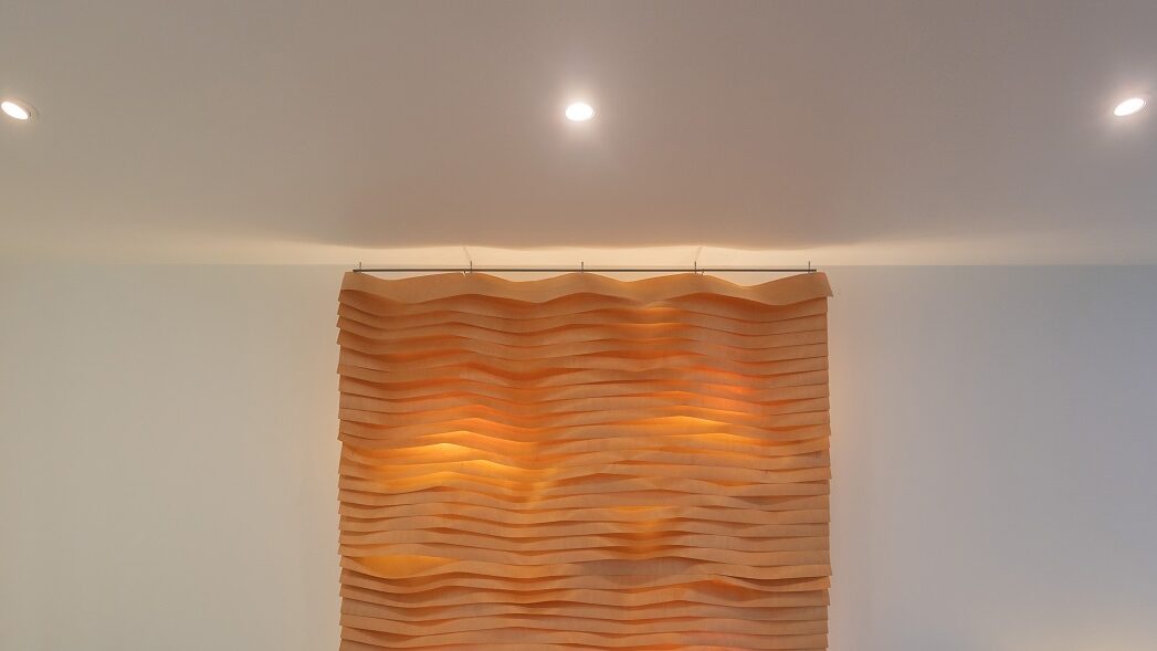 Aaltoileva puupaneelitaulu, jonka lämmin LED-valaistus korostaa pintaa, asennettu valkoiselle seinälle alasvaloilla.