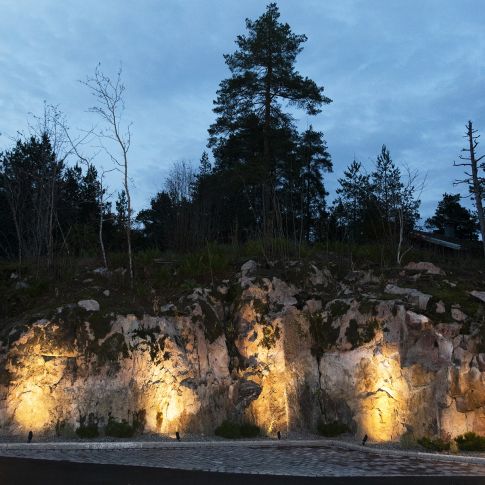 image - LedStore Kuvittele kallioinen mäki, jolla on useita puita, jotka piirtyvät siluettina pilvistä iltataivasta vasten. LedStore.fi:n maahan asennettavat LED-valot valaisevat kalliota, luoden lämpimiä keltaisia hehkuja karhealle pinnalle. Tunnelma on rauhallinen ja luonnollinen.