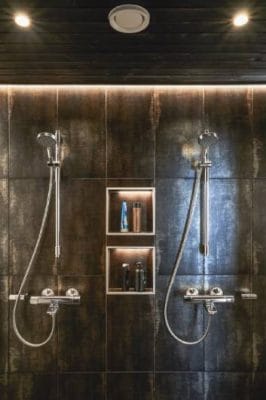 Modern shower area with dual rainfall heads, handheld wands on dark tiled wall; recessed shelf with toiletries under soft, sleek LED lighting.