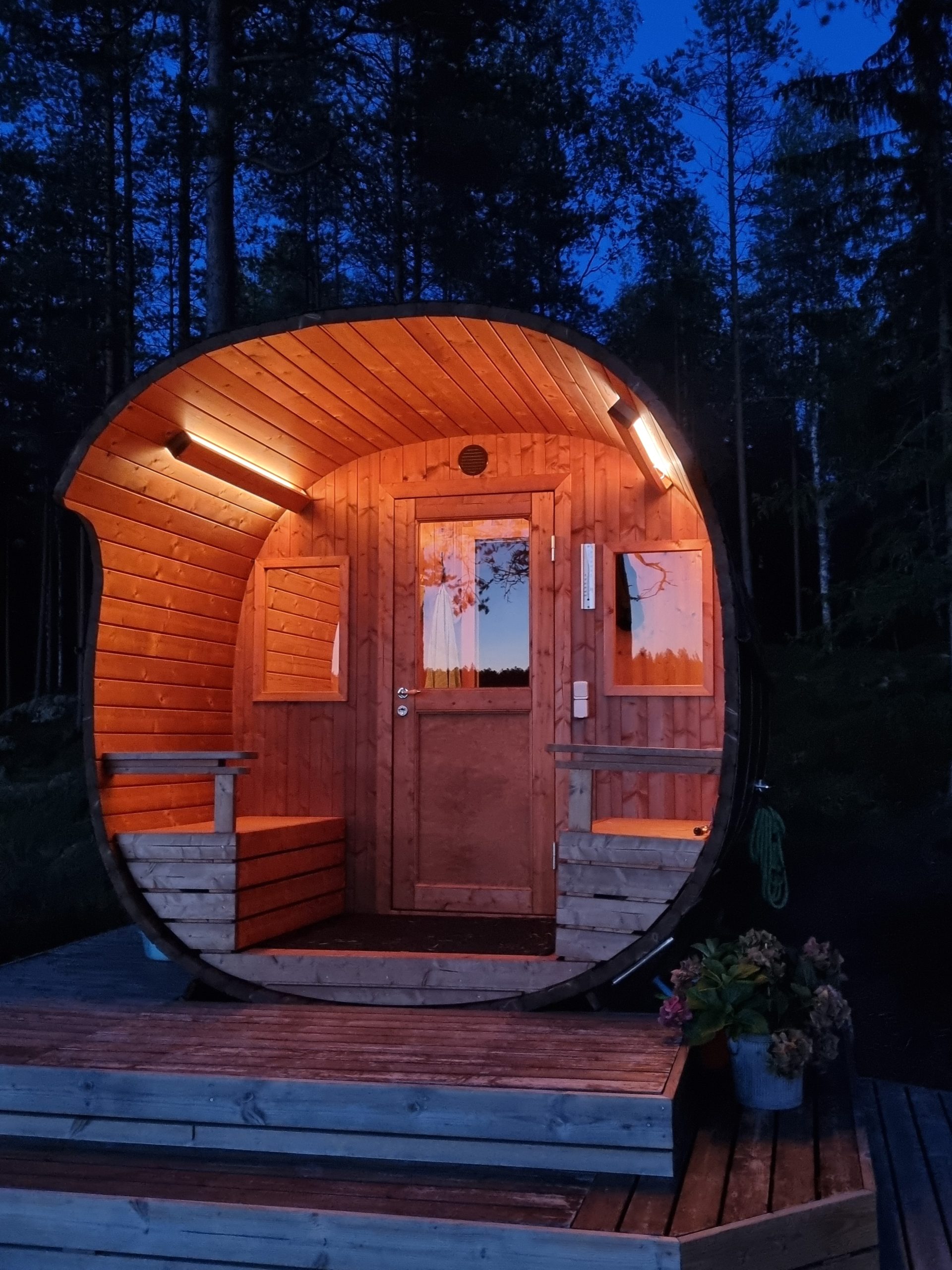 Sauna at dusk - LedStore A small, curved sauna glows warmly in the evening twilight, with LED lights from LedStore.fi highlighting the ambiance of the space. It is situated on a wooden terrace, surrounded by trees, and the colors of flowers in pots add to the charm of this tranquil setting.
