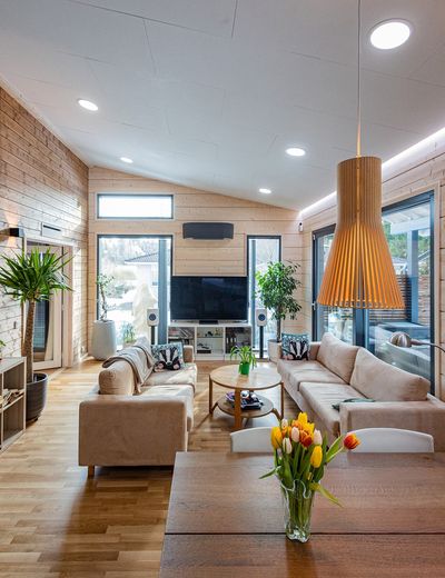 Modern living room with large windows, beige sofas, TV, plants, wooden table with tulip vases, wooden panel walls - illuminated with stylish LED lighting.