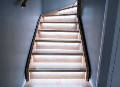 A modern wooden staircase with LED lights under each step, highlighting the minimalist design with warm lighting against grey walls.