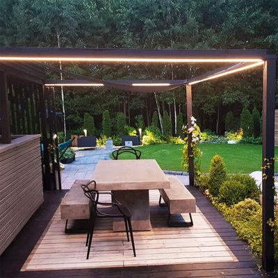 Modern patio with concrete seats, wooden deck and pergola with LED lights; lush garden and trees in the background.