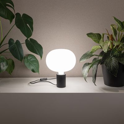 Modern LED table lamp with white round shade on shelf, surrounded by green potted plants for elegant home lighting.