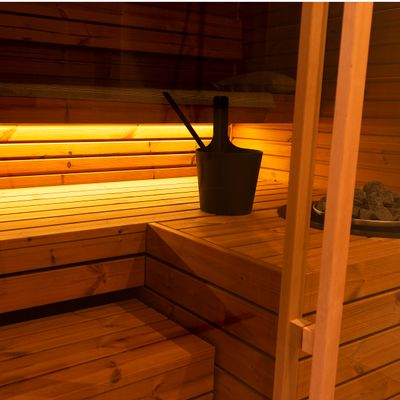 Wooden sauna interior with benches, soft LED lighting, black bucket and scoop on the bench for a cosy and relaxing atmosphere.