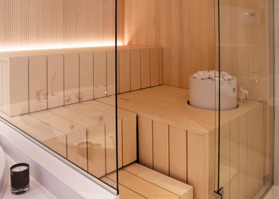 Modern sauna room with stylish light wood benches, glass walls and a bucket of white stones next to the LED lighting next to the stove.