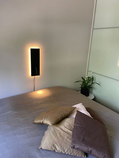 A cosy bedroom with a neatly made bed, cushions, a backlit LED wall light and a plant on the bedside table for a modern feel.