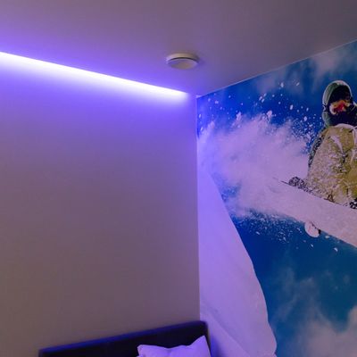 Room with white walls, purple LED ceiling light, skier painting on the wall; visible pillow and part of the bed. Modern and cosy bedroom lighting solution.