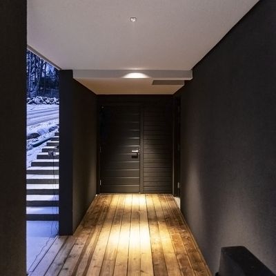 Modern entrance hall with wooden floor, dark walls, soft LED lighting creates atmosphere; black door; snow-covered stairs visible outside.