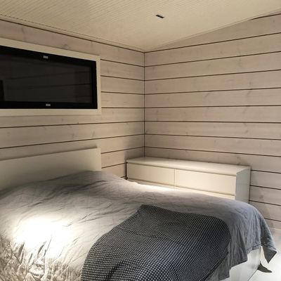 A cosy bedroom with wood panelled walls, atmospheric LED lighting, grey bed linen on the bed and a white chest of drawers under a small window to create a peaceful atmosphere.