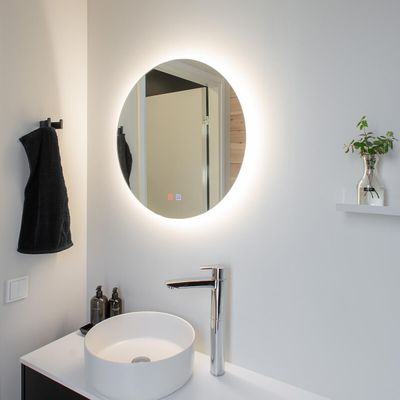 Minimalist bathroom with LED illuminated round mirror, white sink with chrome tap, black towel and small plant on shelf.