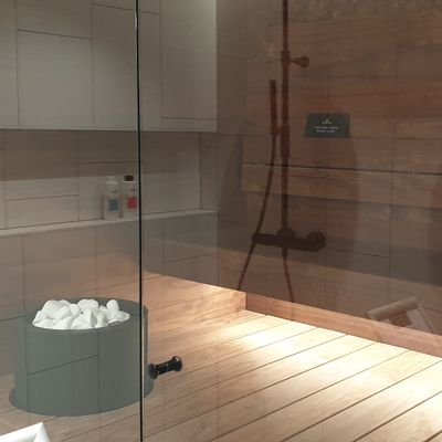 Modern sauna room with wooden walls, glass door, stone bucket, shower head; perfect atmosphere with LED lighting for relaxation and comfort.
