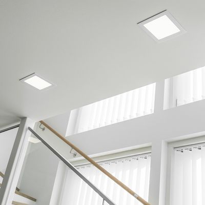 Bright square recessed ceiling lights illuminate a modern staircase with glass balustrade and large windows with vertical curtains.