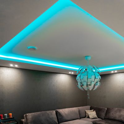 A modern living room with a grey sofa, blue-green LED ceiling lights, a circular pendant light and a small table with vivid candles for ambient lighting.