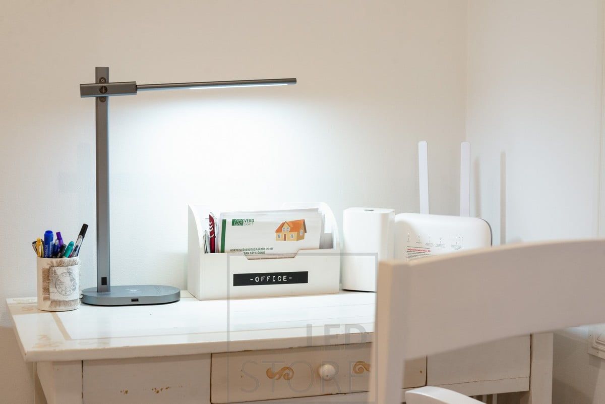 A crane table lamp illuminating a workstation. Led table lamp KURKI bedside table lamp and reading lamp in one. KURKI is a metal frame, grey, simple and elegant lamp. The KURKI led table lamp provides light directly or indirectly thanks to the freely adjustable body. Wireless and USB charging for your phone. Colour temperature adjustable and dimmable. Ledstore.fi