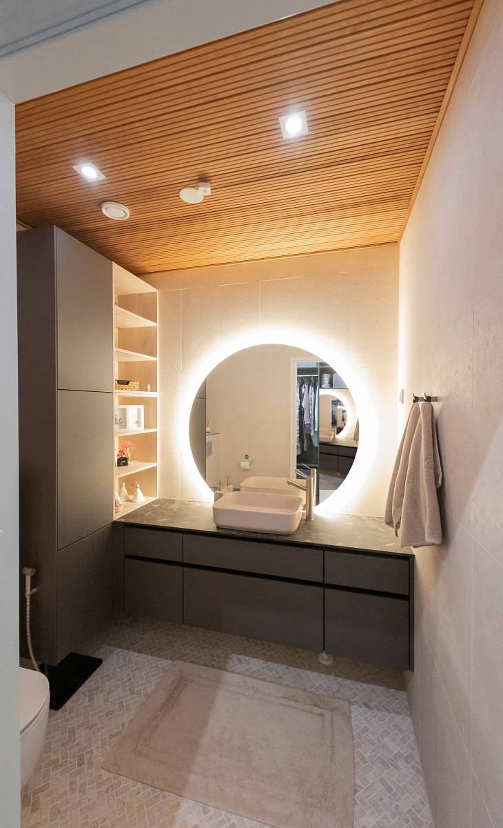 A modern bathroom with LED lighting from LedStore.fi: a circular LED backlit mirror above the sink creates an elegant atmosphere. The bathroom features dark cabinets, warm wooden ceilings and light walls. Towels are placed on the right side and shelves await you on the left. A beige carpet rests on the floor, adding to the overall effect.