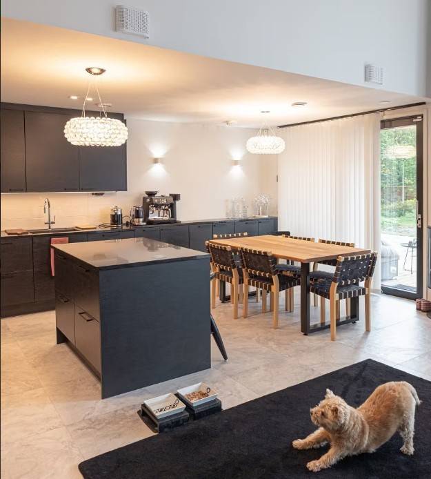 A modern kitchen and dining area with black cabinets, a wooden dining table and comfortable LedStore.fi LED lighting create a cosy atmosphere. A cat stretching on a black carpet observes the cosy ensemble. 