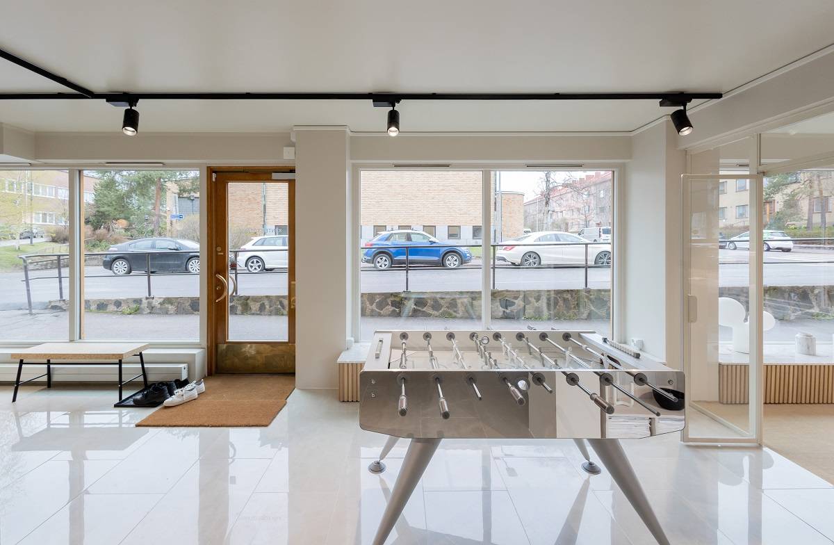Simple and efficient office lighting for Kiinteistötakuu A modern interior with large windows showcasing cars parked outside, with a football table on the shiny floor. A bench with a shoe rack and door mat sits around under LED track lighting, highlighting the modern design of the room. All this can be found in LedStore.fi's wide range of LED lighting solutions for your home interior.