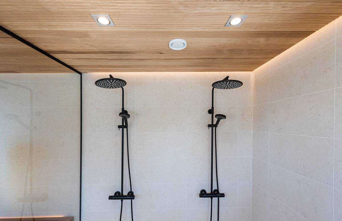 The modern shower room has two black shower heads for cream-coloured tiling. The wood panelled ceiling, accented by recessed LED lights from LedStore.fi, creates a minimalist and elegant look. The shower area is separated from the rest of the space by a glass exterior wall.  