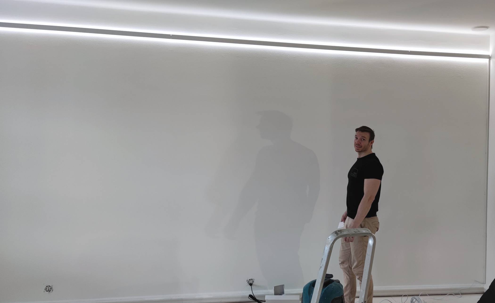 A person on a ladder with a tool, in soft LED lighting from above, against a white wall. Cables and equipment scattered on the floor in LED shop fittings.