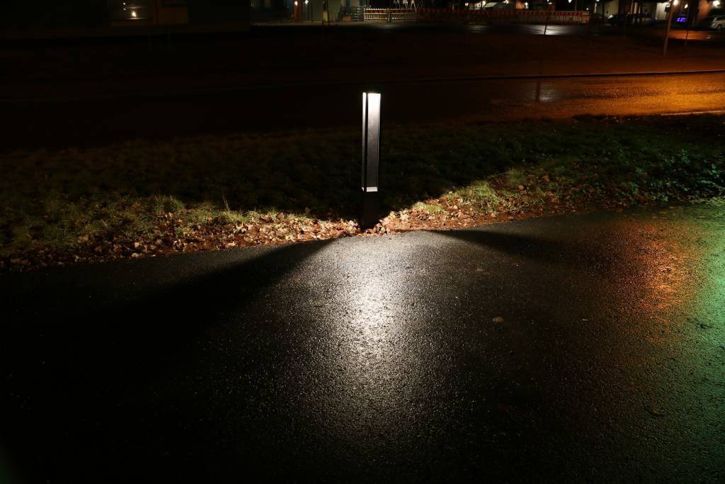 Dimly lit street at night with an LED ground light casting shadows on wet pavement and grass; blurred background with subtle dim lighting.